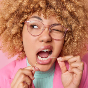 Imagem de uma jovem com cabelos cacheados e amarelos, usando fio dental pisca os olhos e cuida da higiene bucal. Usa óculos transparentes roupas elegantes isoladas sobre fundo rosa.